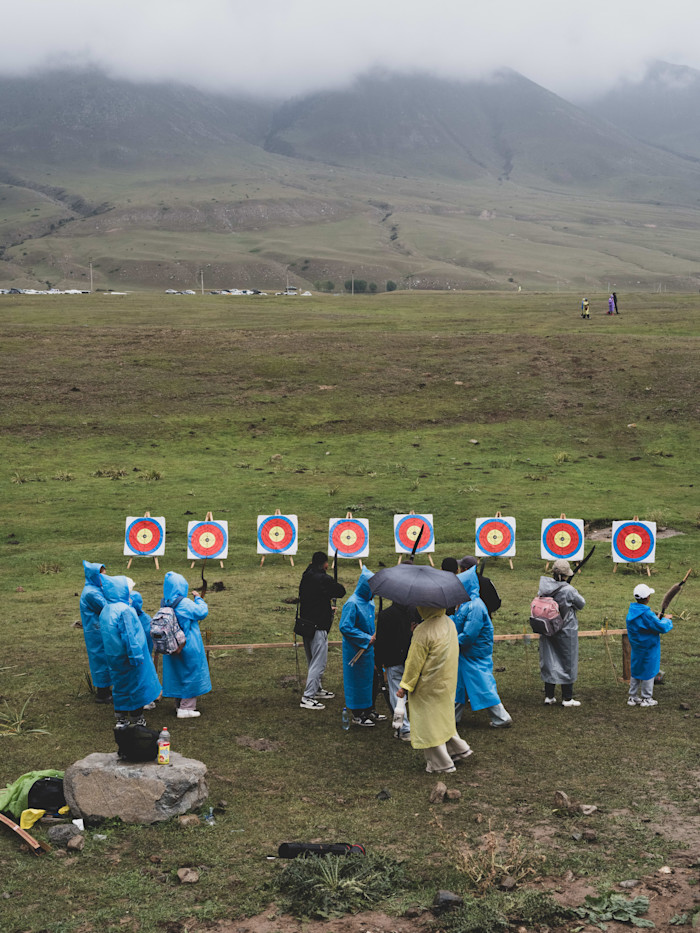 People in raincoats stand in front of archery targets on a grassy field, with mountains in the background.