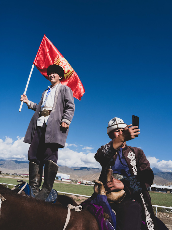 A man stands on horseback holding the Kyrgyz flag, while another man beside him takes a selfie with his phone. Both wear traditional clothing.