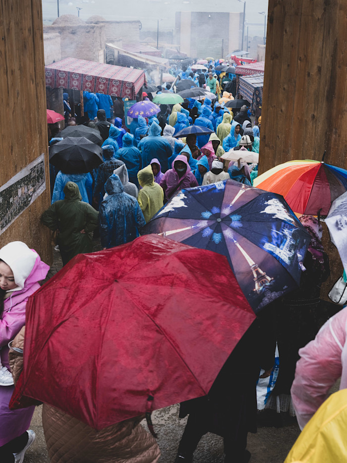 Crowd of people in colorful raincoats and umbrellas gather in a food area at the World Nomad Games despite rainy weather.