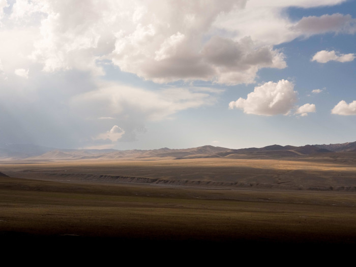 Vast, open landscape with rolling hills under a dramatic sky filled with scattered clouds and sunlight casting shadows over the plains.