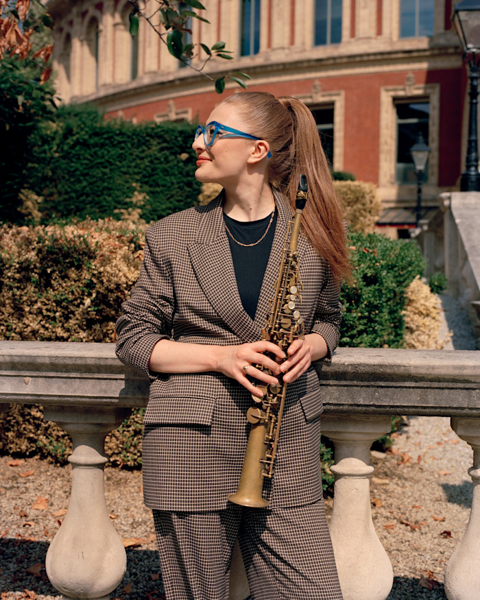 Jess Gillam stands in front of the Royal Albert Hall in a checked suit, holding a saxophone and looking to the side.