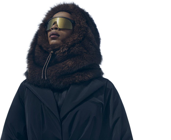 A model wearing a dark ski jacket with a fur-trimmed hood and reflective goggles.