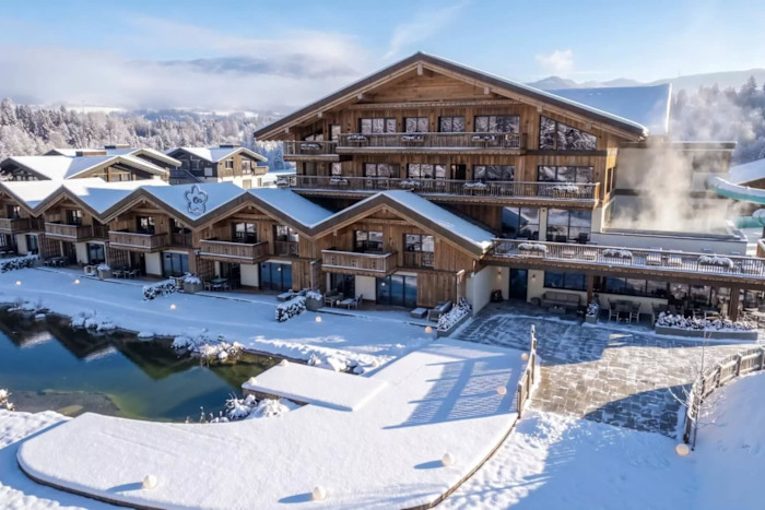 A large chalet-style hotel with wooden exterior, snow-covered roofs and balconies overlooks a small pond and snowy landscape. Steam rises from an outdoor area