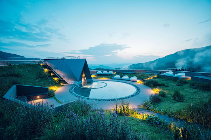 A rooftop terrace with landscaped gardens, a circular reflecting pool, modern geometric structures and mountain views at dusk