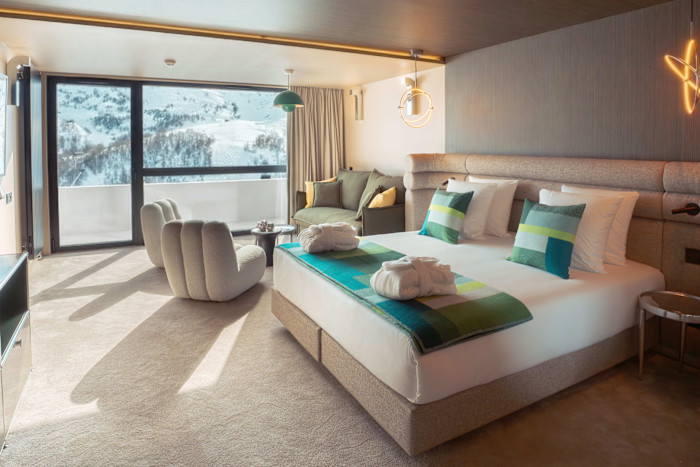 Modern hotel room with a large bed, two plush chairs, a small sofa and floor-to-ceiling windows overlooking snowy mountains