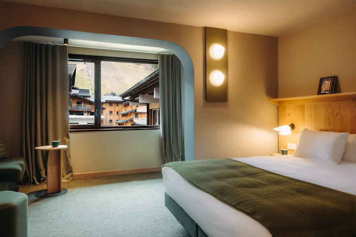 A cosy hotel room with a large window overlooking wooden chalet buildings and a grassy ski slope. The room features a neatly made bed with white linens and a green throw, a small round table with a mug and warm lighting