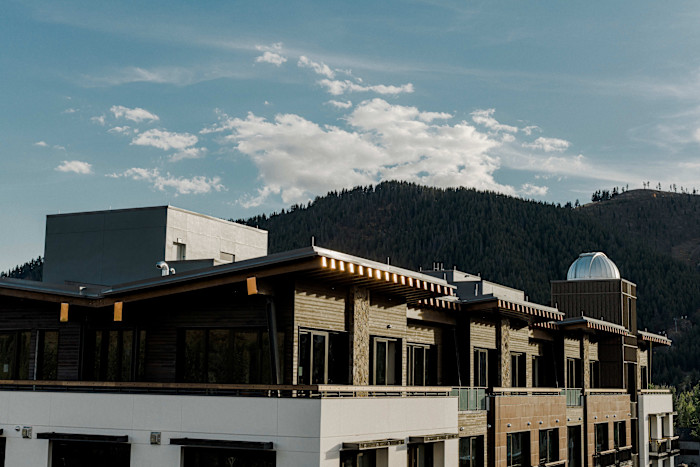 A modern building with wood and stone accents, featuring a dome observatory on the rooftop, set against forested hills and a partly cloudy sky