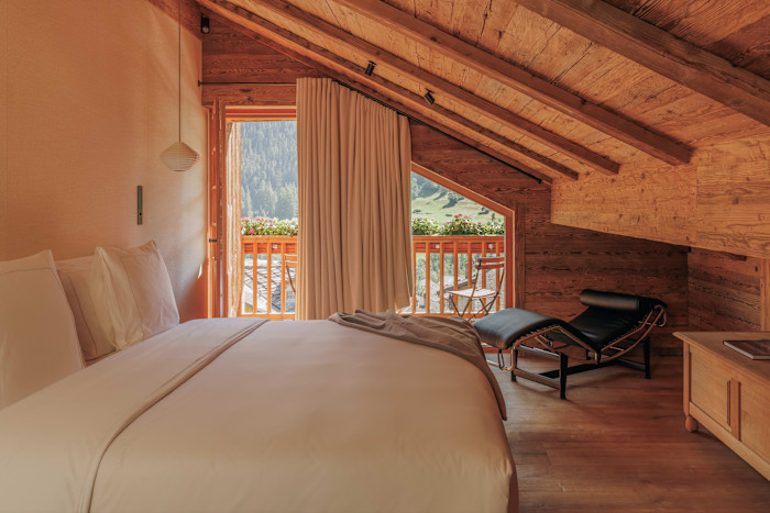 A cosy hotel room with a sloped wooden ceiling, large bed, lounge chair and a balcony overlooking a green mountain landscape