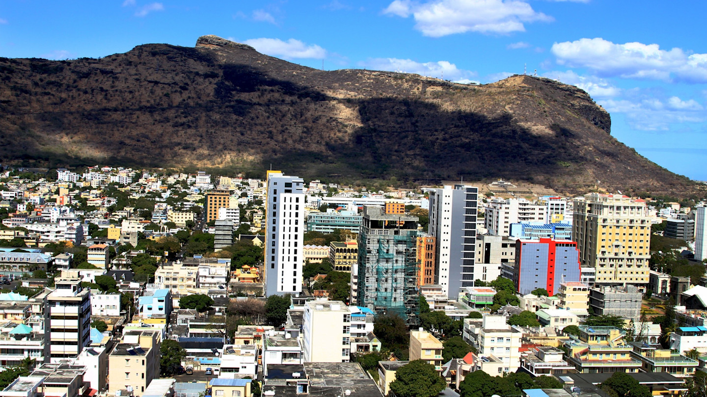 Skyline of Port Louis with modern high-rise buildings in the foreground and a large mountain in the background under a blue sky.