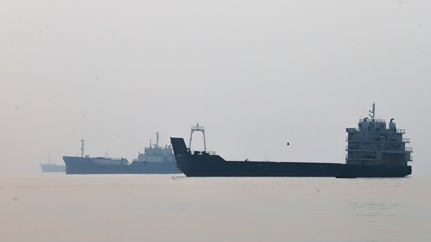 Several large commercial vessels are anchored in calm waters under a hazy sky in the Strait of Hormuz.