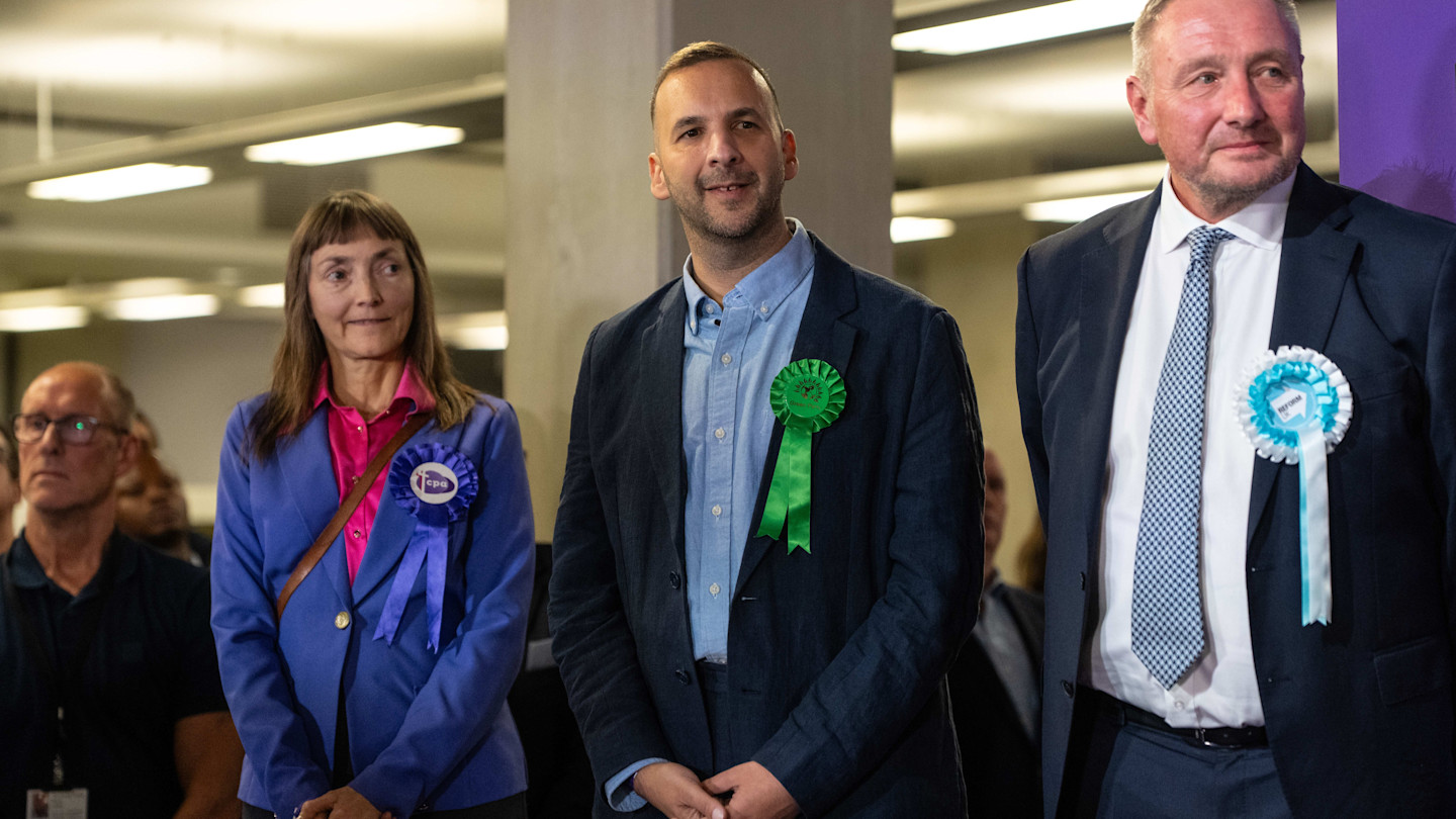 Zack Polanski, wearing a Green Party rosette, stands between two other candidates at the Holborn and St Pancras by-election count.