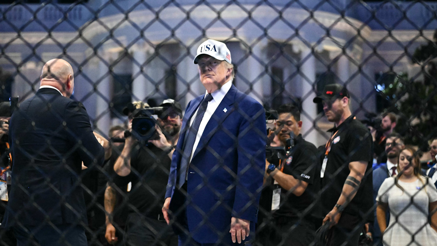 Donald Trump stands in the center of an MMA cage wearing a suit and a “USA” hat, surrounded by cameramen and event staff.