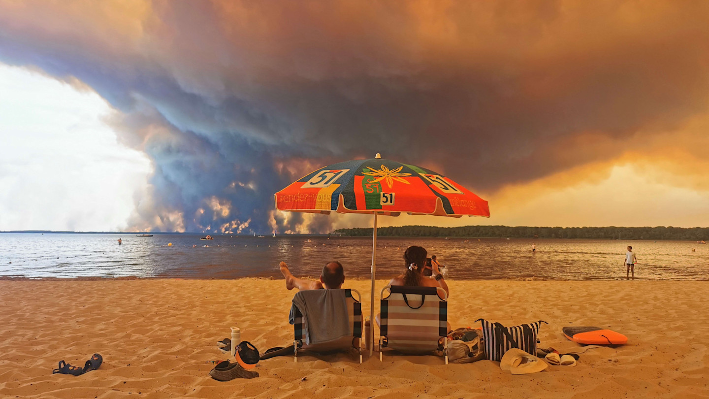 Beachgoers are seen lounging under a colourful umbrella on a beach as thick smoke from a wildfire rises dramatically across the water in the distance.