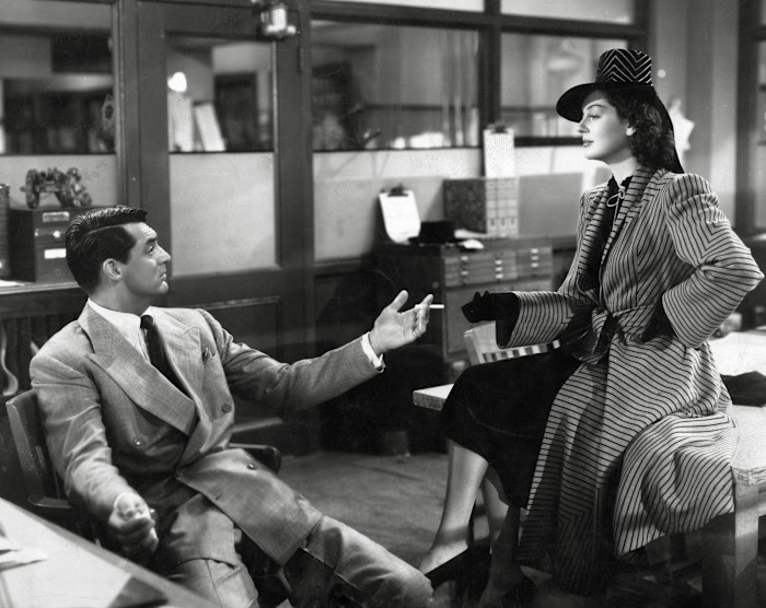 Cary Grant and Rosalind Russell in an office scene from "His Girl Friday". Grant gestures while Russell sits on a desk in a striped coat and hat.