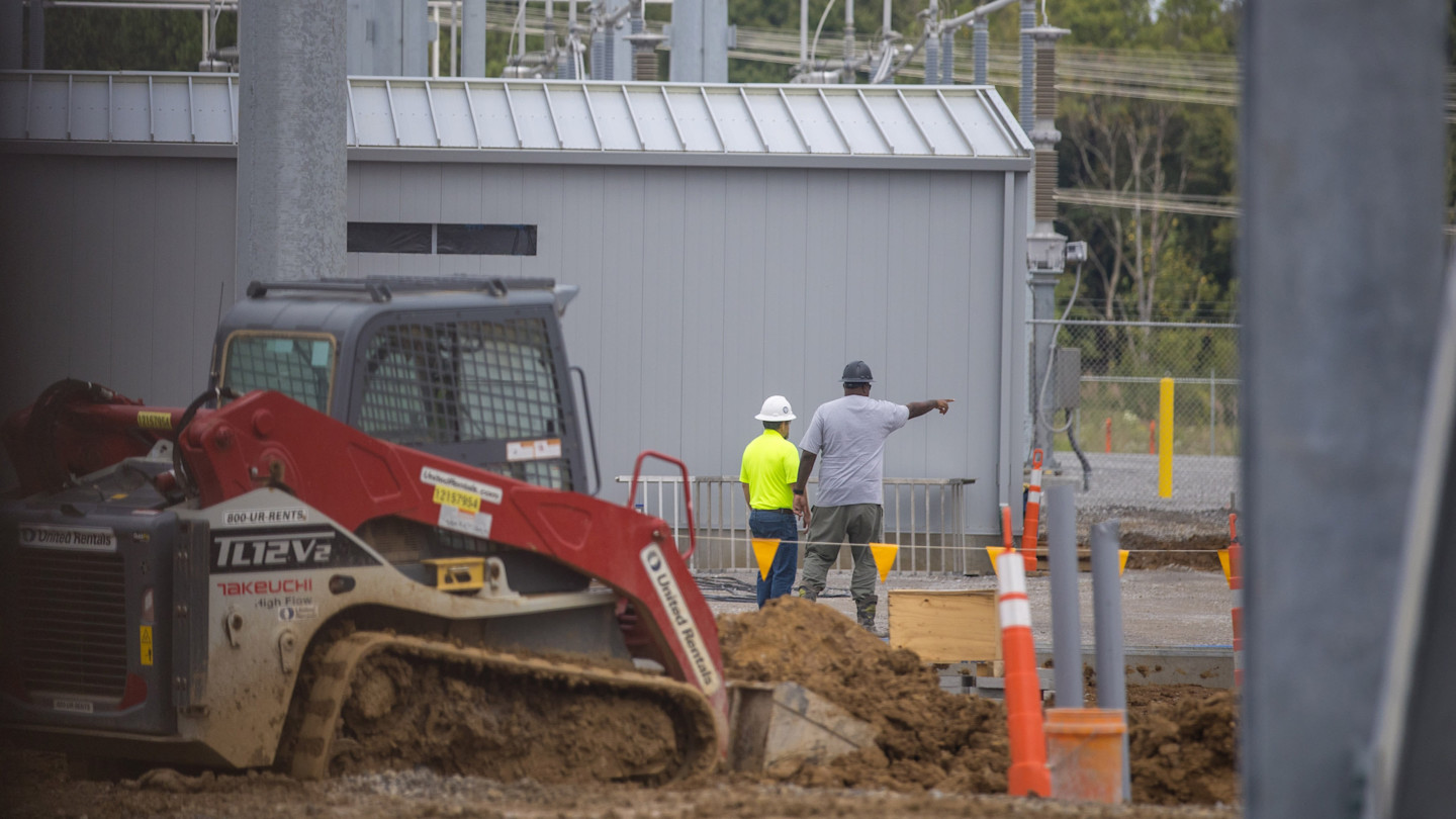 Two construction workers wearing hard hats stand near a building at the River Bend AI data centre site, with one pointing. A muddy skid steer loader is in the foreground.