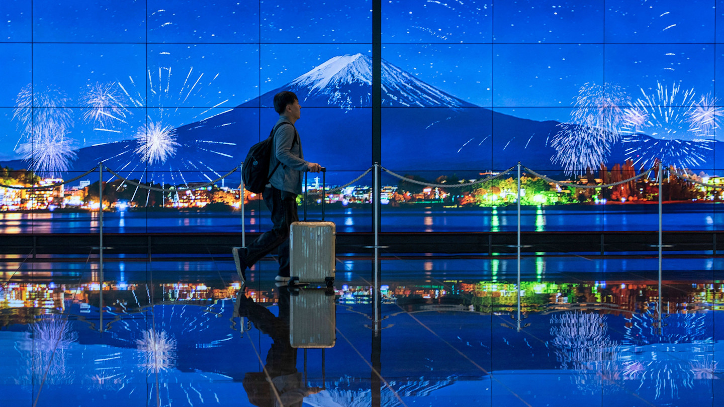 A traveller with a suitcase walks past a large digital display of Mount Fuji and fireworks at Haneda Airport arrivals terminal.