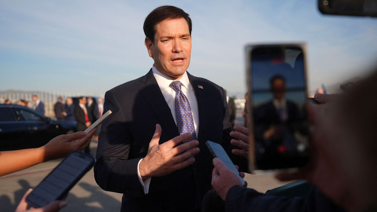 Marco Rubio speaks to reporters outside, gesturing with his hands, as microphones and phones are held towards him.