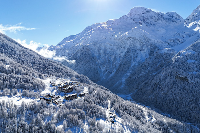 A snow-covered alpine village surrounded by dense forest with tall mountains in the background under a clear blue sky.