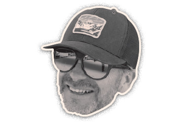 Black-and-white cut-out portrait of a smiling man wearing a baseball cap and glasses