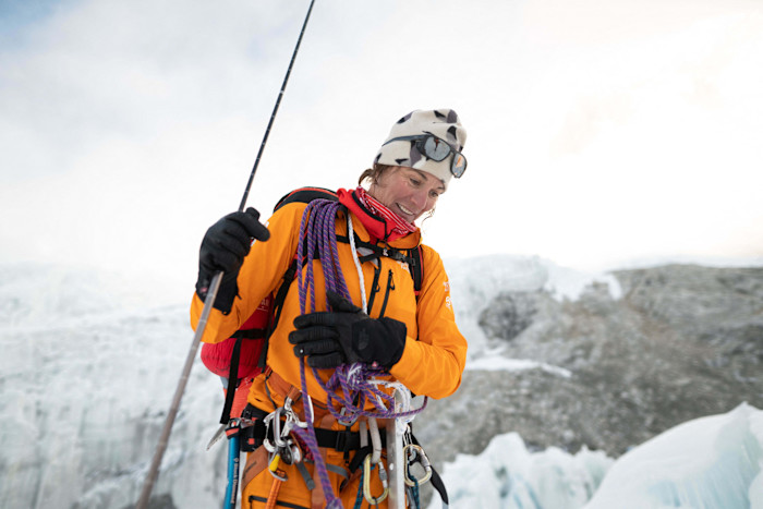 Hilaree Nelson stands on a snowy mountain, smiling and holding climbing rope, wearing an orange jacket and mountaineering gear.