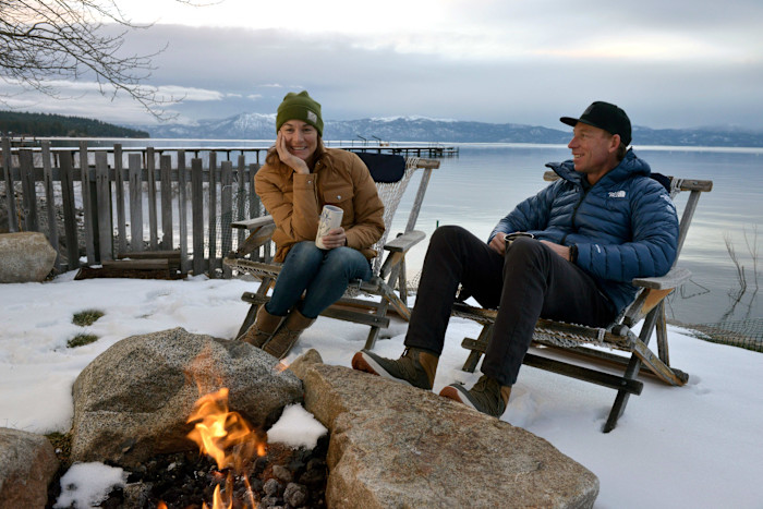 Jim Morrison and Hilaree Nelson sit in chairs by a fire pit on snow, overlooking Lake Tahoe with mountains in the background.