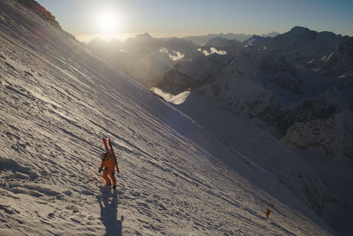 Jim Morrison, wearing an orange suit and carrying skis, climbs a steep snowy slope with a team member following behind at sunrise.