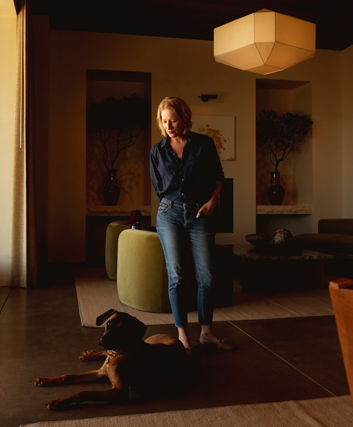 A woman in a navy shirt and jeans stands beside a brown dog lying on the floor of a warmly lit living room with contemporary furnishings.
