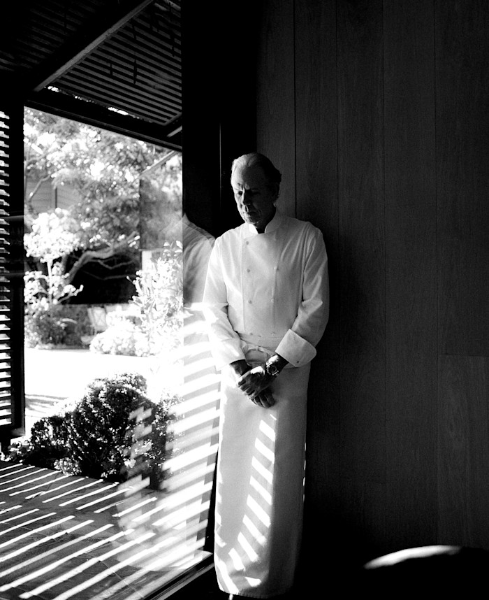 Black-and-white portrait of a chef in a white jacket and apron standing beside a large window, with striped sunlight falling across his clothing and a garden visible outside.