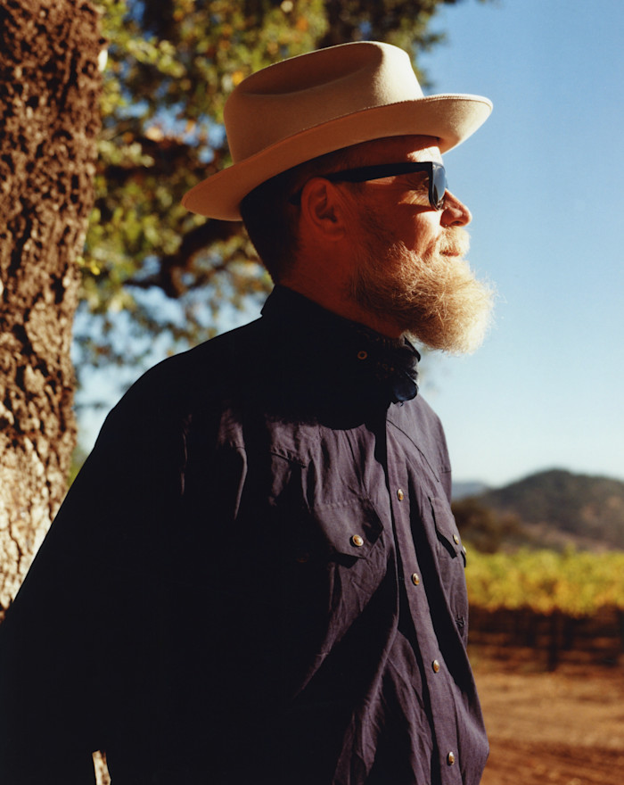 Side-profile portrait of a bearded man wearing a cream-coloured wide-brimmed hat, dark sunglasses and a navy shirt, standing in bright sunlight beside a tree, with a vineyard and hills behind him.