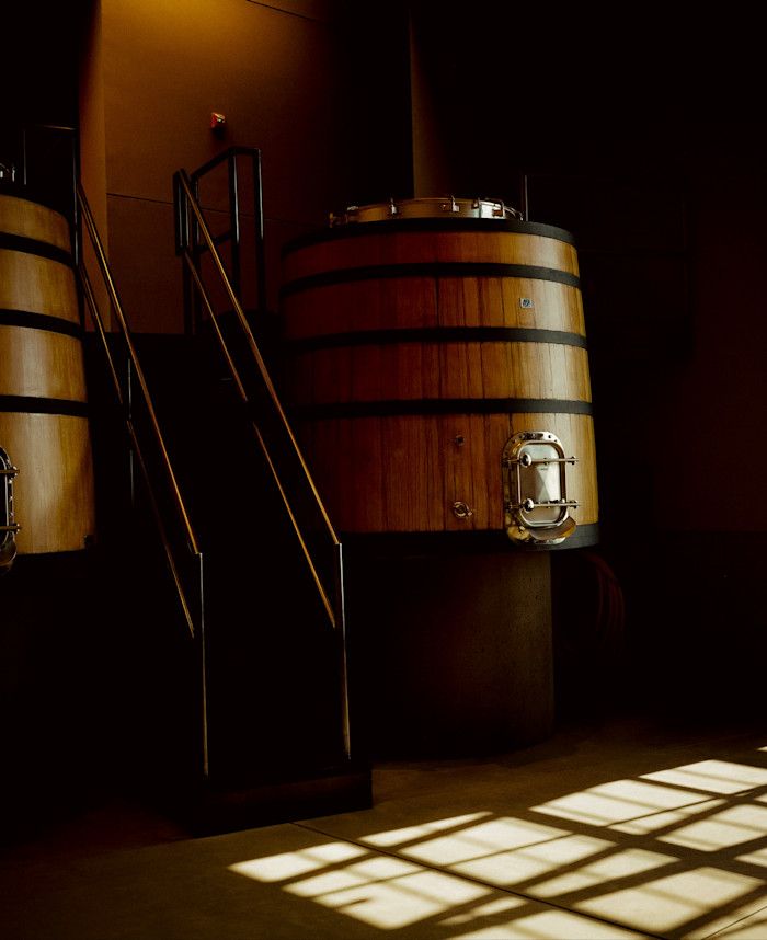 Large cylindrical wooden wine fermentation vats with dark metal bands stand inside a dimly lit winery, with a metal staircase between them and sunlight casting geometric shadows across the floor.