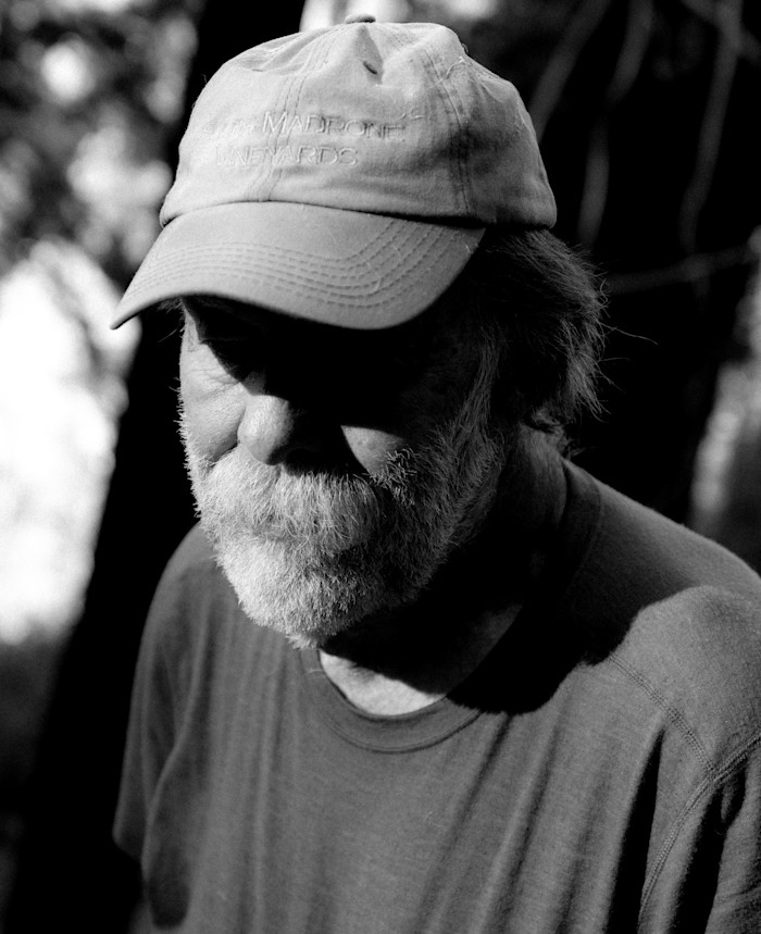 Black-and-white close-up portrait of a bearded man wearing a light-coloured baseball cap and T-shirt, looking down, with sunlight illuminating part of his face.