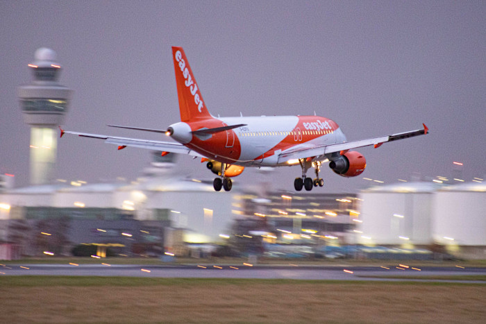 EasyJet Airbus A319 with registration G-EZFU landing at Amsterdam Schiphol Airport, with the control tower and terminal in the background.