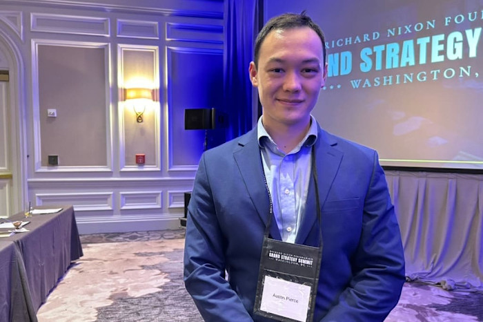 Austin Pierce stands in a conference room at the Grand Strategy Summit, wearing a suit and summit name badge.