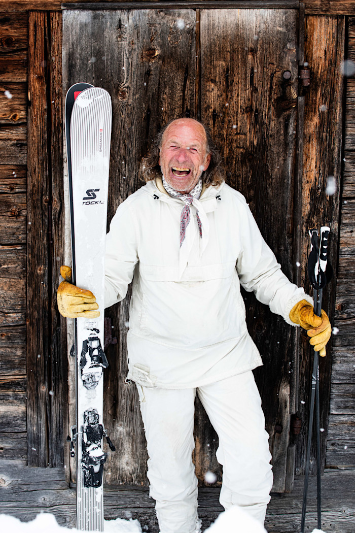 Heinrich Giesker, dressed in white ski gear and yellow gloves, smiles while holding skis and poles in front of a wooden wall in snowy Engelberg.