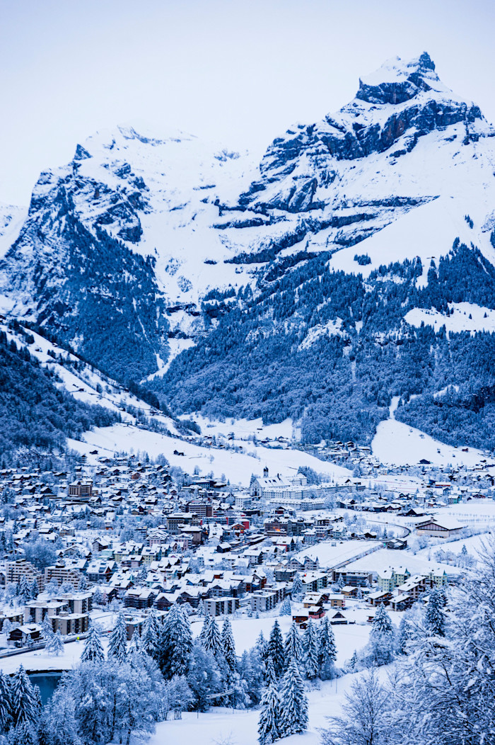 A snow-covered town with clustered buildings and evergreen trees sits below towering, rugged mountains in Engelberg, Switzerland.
