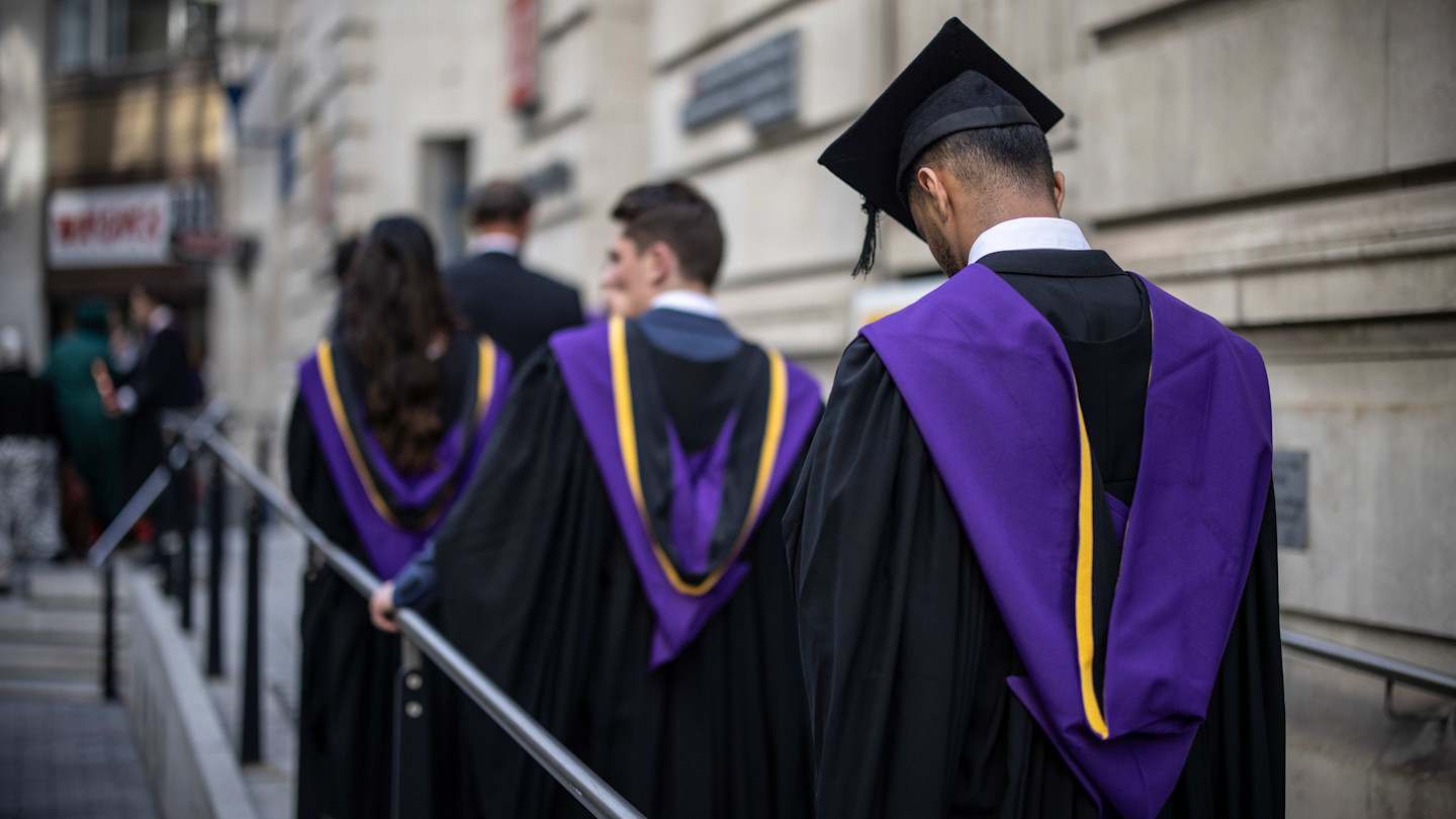 Students in graduation gowns and caps with purple and yellow hoods walk in a line outside a building.