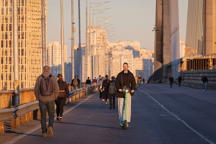 People cross a bridge on foot and by scooter in Kyiv during an air raid alert, with damaged infrastructure visible and traffic disrupted.