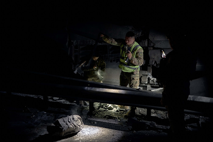 Emergency workers in military uniforms examine damage on a bridge at night, shining flashlights on debris.