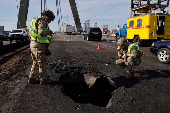 Two police officers in military gear inspect a large crater in the road on the North Bridge in Kyiv after a Russian drone strike.