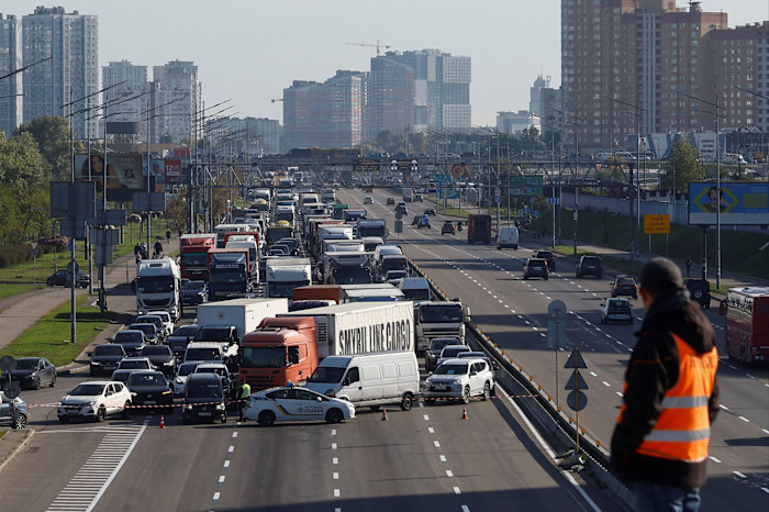 A construction worker in an orange vest looks at a long traffic jam on a multi-lane road, with vehicles blocked by police cars near a bridge in Kyiv.