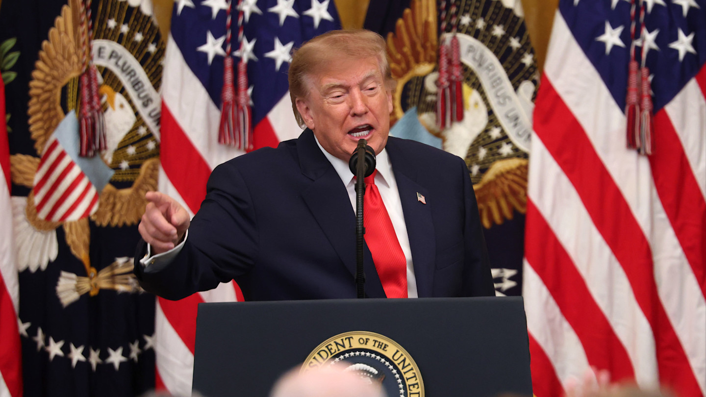 Donald Trump speaks at a podium with the presidential seal, gesturing with his hand, in front of U.S. flags and official emblems.