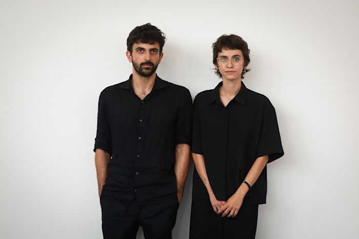 A young man with dark hair and beard stands alongside a bespectacled young woman, both in black clothes and looking at the camera with serious facial expressions.