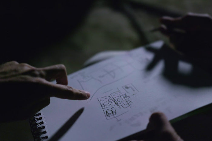 A darkly lit film still of hands pointing at a piece of paper with hand-drawn plans on it.
