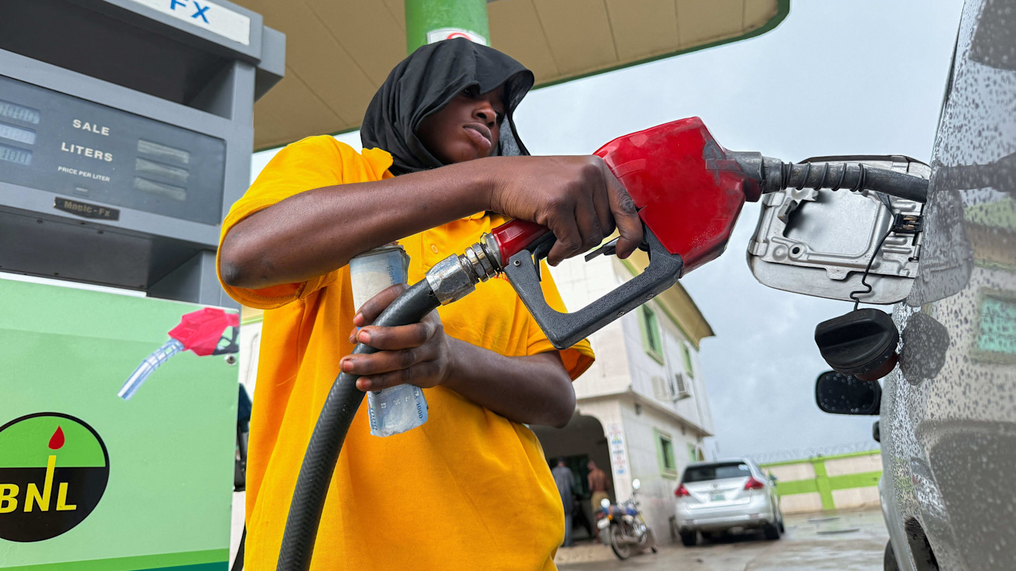 A fuel attendant in a yellow shirt fills a car’s tank with a red pump nozzle at a petrol station.