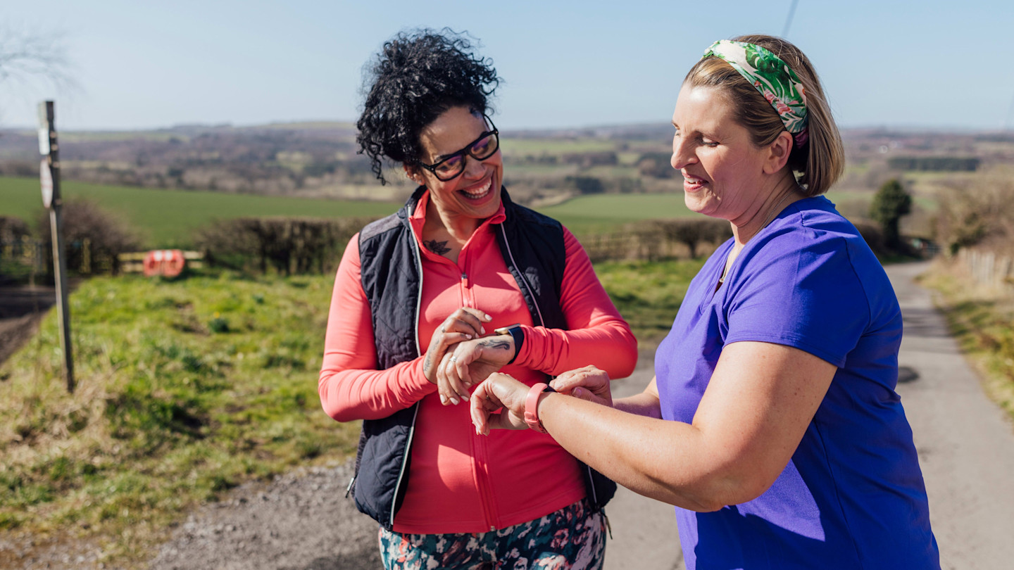 Two women in gym clothes pause on a rural road, smiling while checking their smartwatches with scenic countryside in the background.