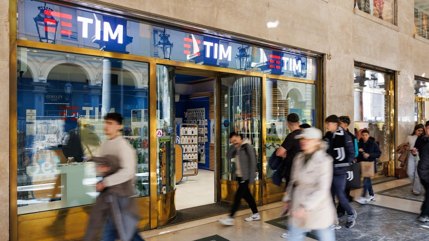 People walk past the entrance of a Telecom Italia (TIM) store in Turin, with store displays visible inside.