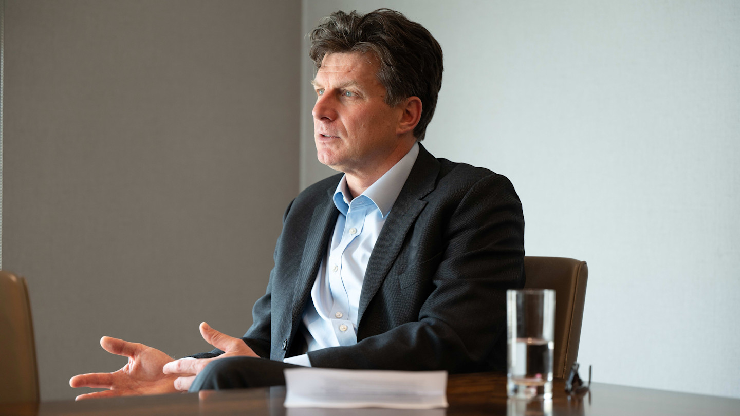Patrick Thomson sits at a table, gesturing with his hands, with a glass of water and papers in front of him.