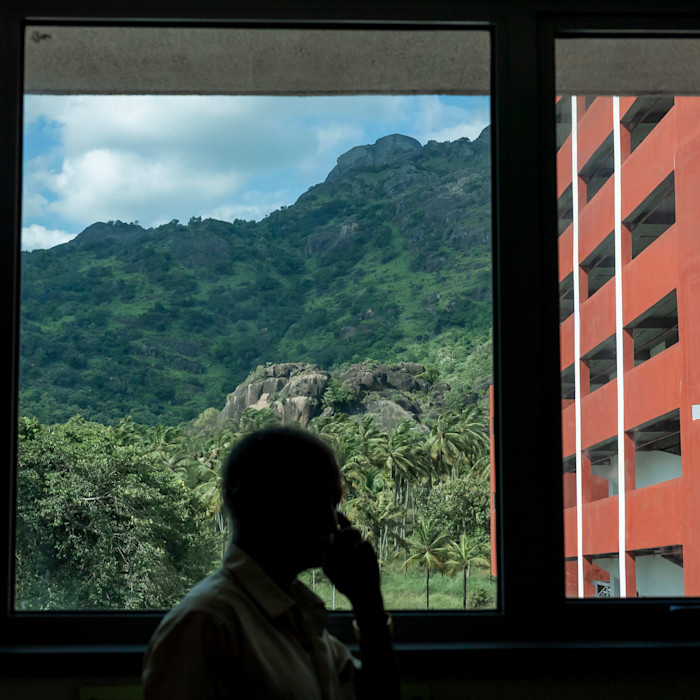 A man in silhouette talks on the phone near a window overlooking green hills and a red building.