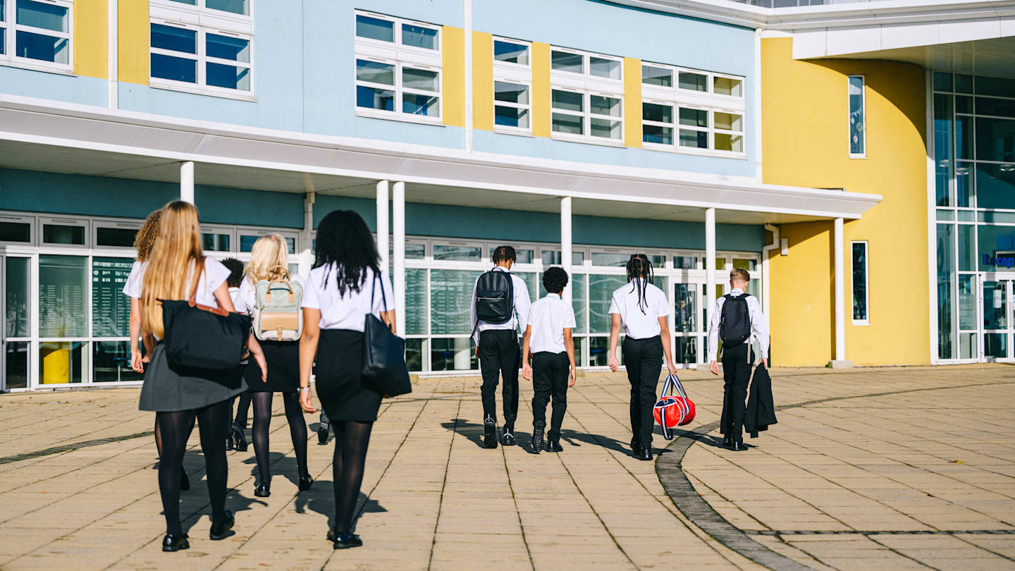 A group of students in school uniforms walk towards the entrance of a modern school building.
