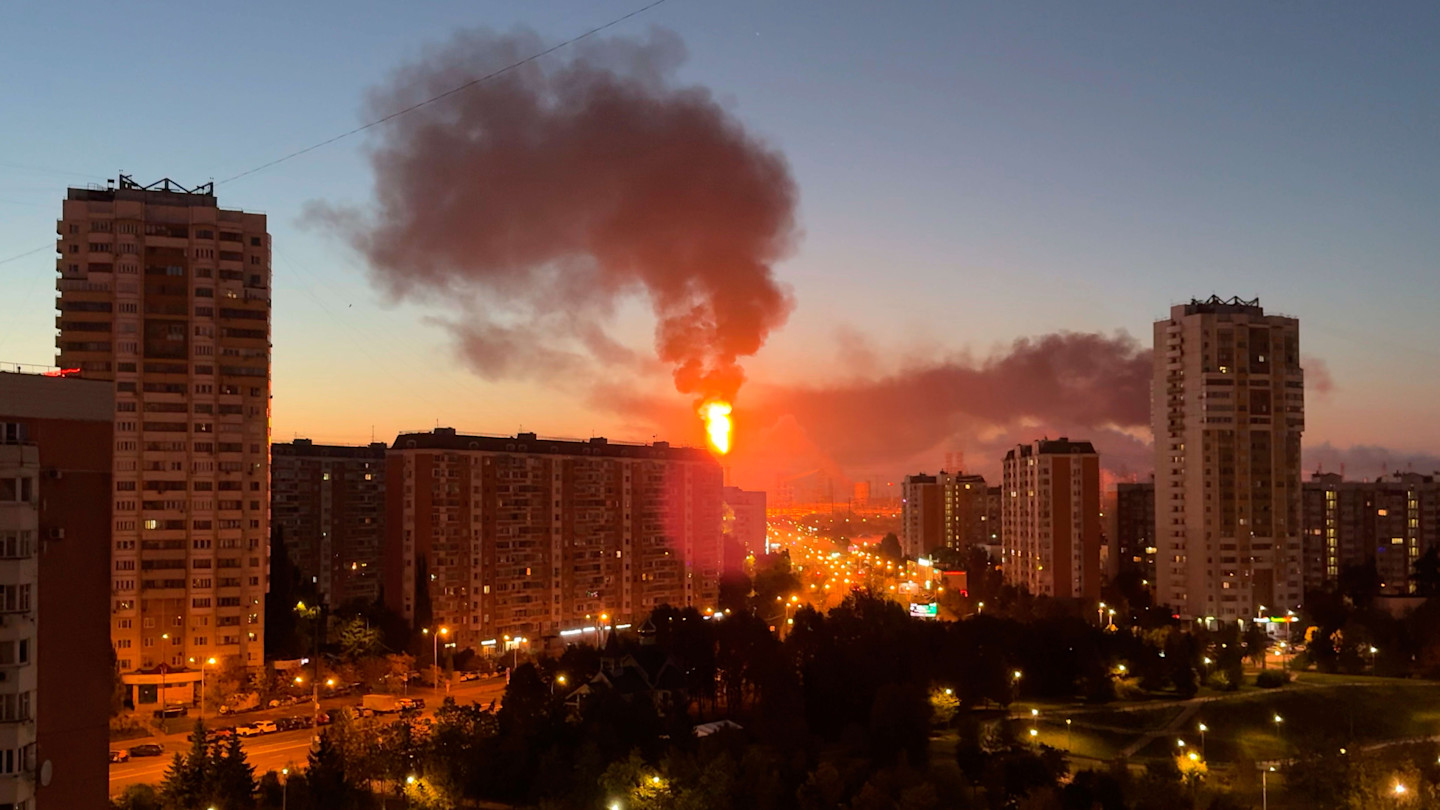 Flames and thick smoke rise from a building behind high-rise apartments in Moscow at dusk.
